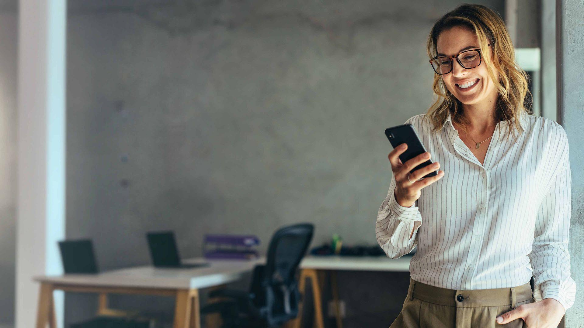 Bild: Frau in weißer Bluse steht in einem Büro, schaut auf Ihr Handy und lächelt.