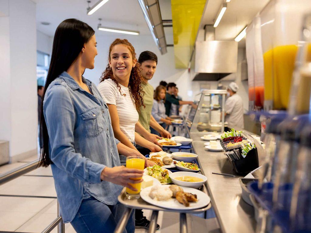 Bild: Menschen stehen in der Kantine mit ihren Tabletts und erhalten Essen.