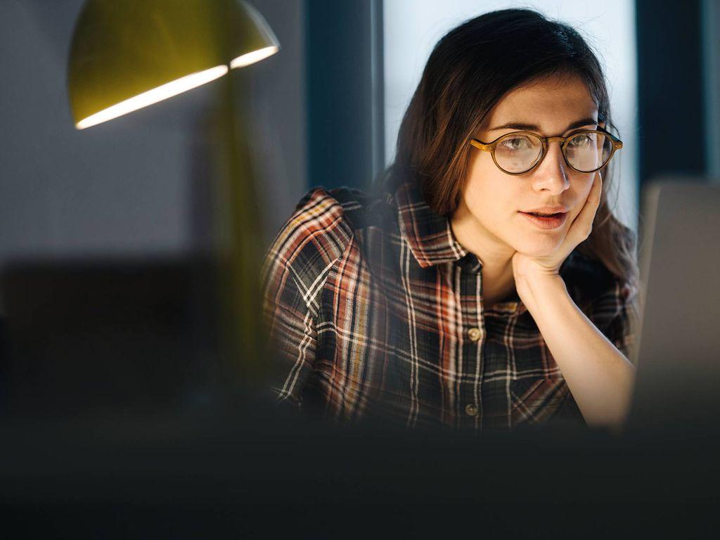 Bild: Junge Frau mit Brille liest etwas auf dem Laptop im Arbeitszimmer.