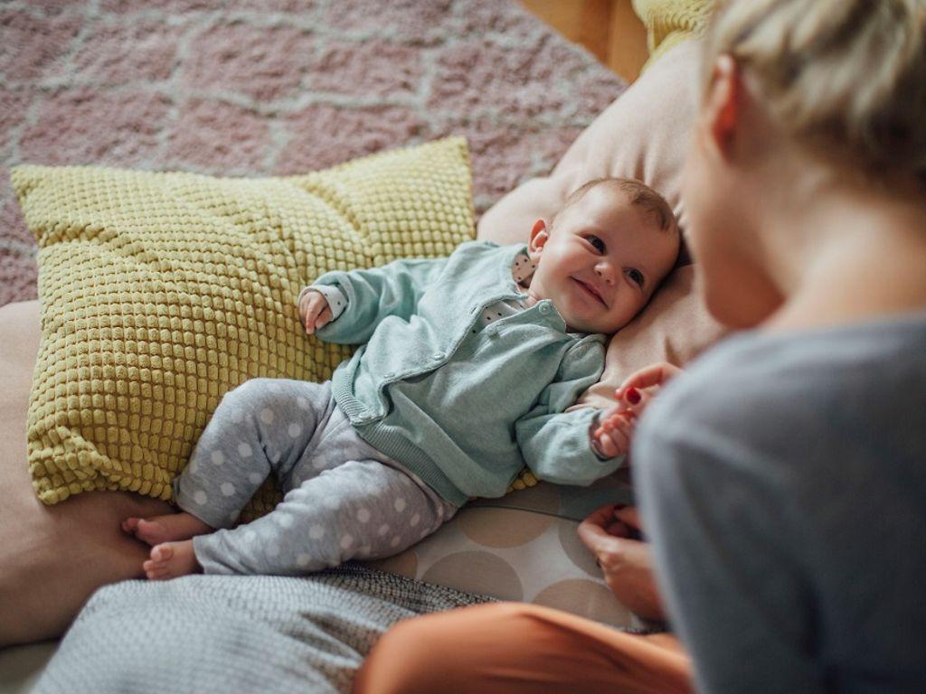 DAK Willkommen Baby: Lächelndes Baby liegt auf Kissen und schaut zu einer Frau hoch.