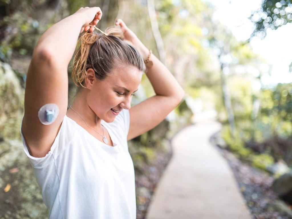 Diabetes: Frau mit Diabetessensor am Arm macht sich einen Zopf in der Natur.