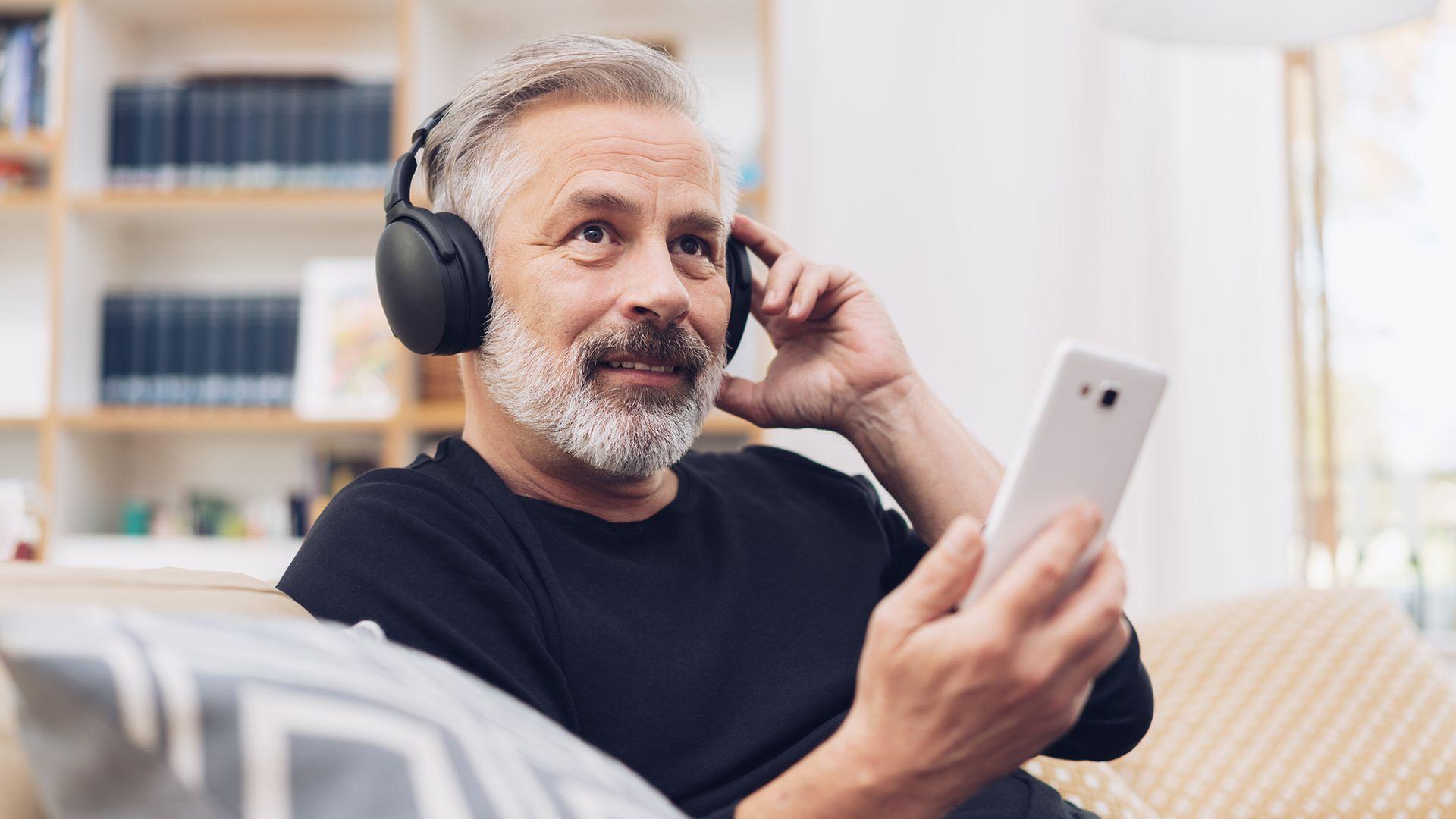 Älterer Mann sitzt auf dem Sofa, hat Kopfhörern auf und sein Handy in der Hand.