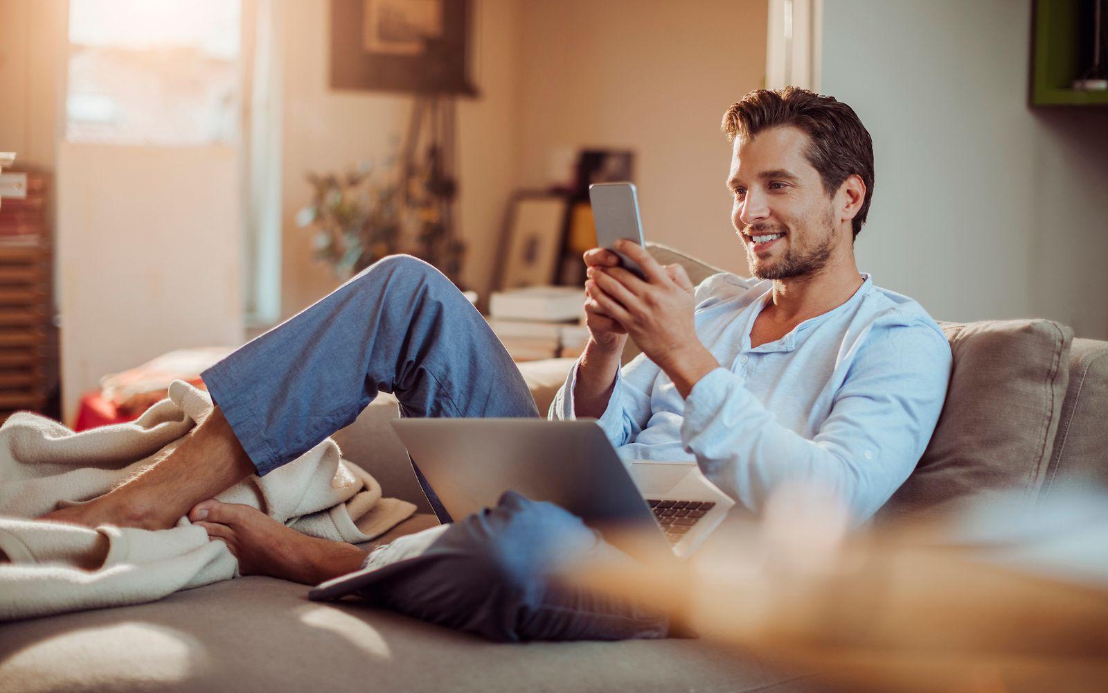 Ein junger Mann sitzt auf dem Sofa mit Handy und Laptop