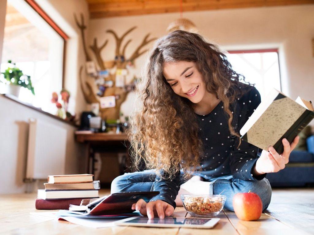 Bild: Jugendliche mit Buch in der Hand sitzt auf dem Boden und bedient ein Tablet.